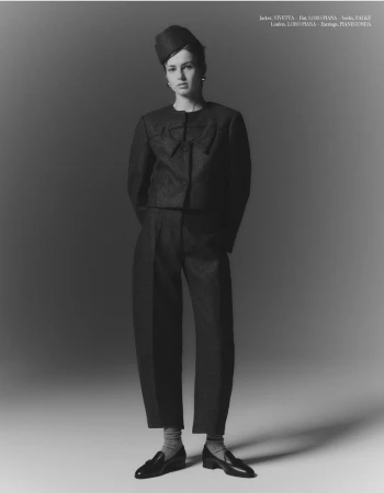 Fashion model poses in a structured, monochromatic suit with a bow-detailed jacket, wide-leg pants, loafers, and a small hat in a studio setting.