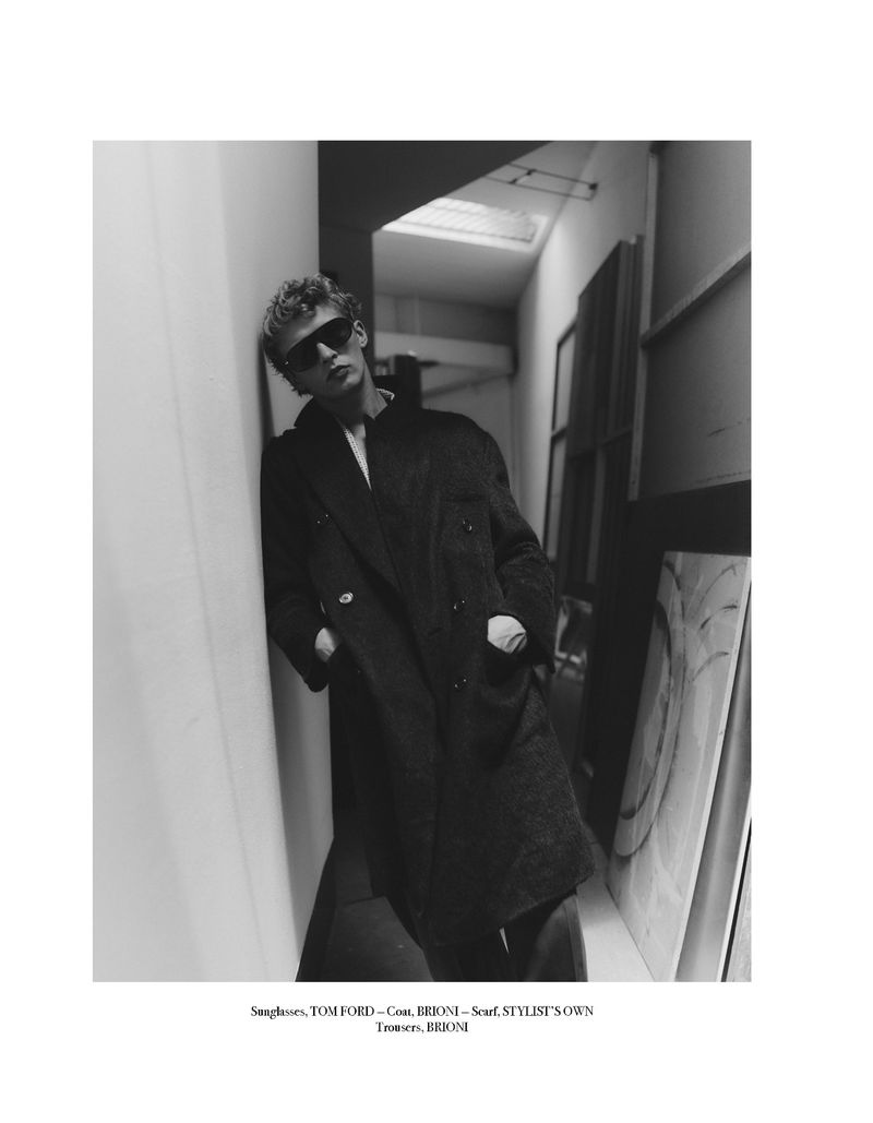 Monochrome portrait of a man with sunglasses in a dark coat leaning against a wall indoors, with artistic prints nearby.