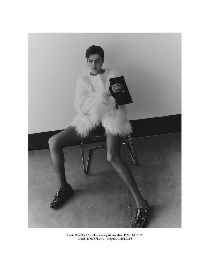 Black and white studio shot of a woman seated in an oversized fur coat, holding a clutch, and wearing loafers.