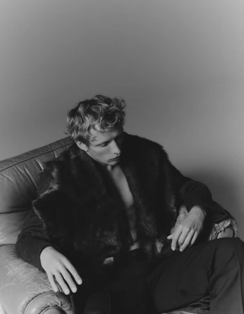 Monochrome portrait of a young adult man with fair hair in a faux fur coat, seated on a leather armchair against a plain grey backdrop.