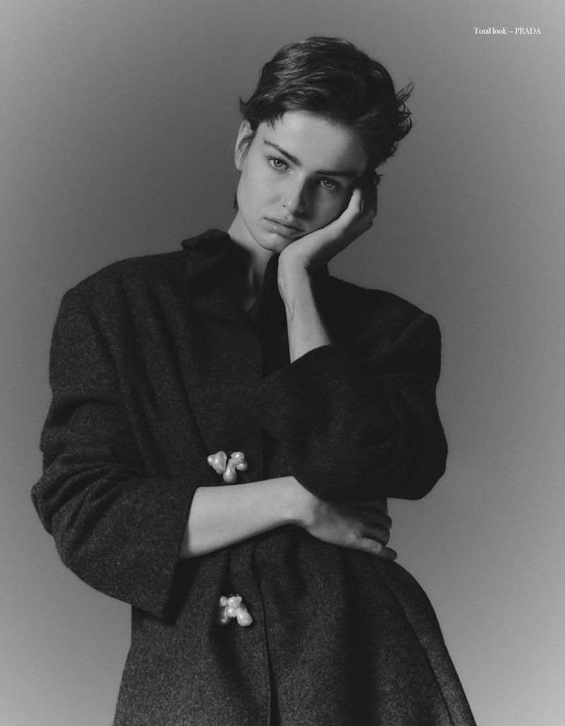 Monochrome portrait of a young woman with short dark hair wearing a dark coat with embellishments, posing against a grey backdrop.