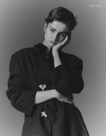 Monochrome portrait of a young woman with short dark hair wearing a dark coat with embellishments, posing against a grey backdrop.