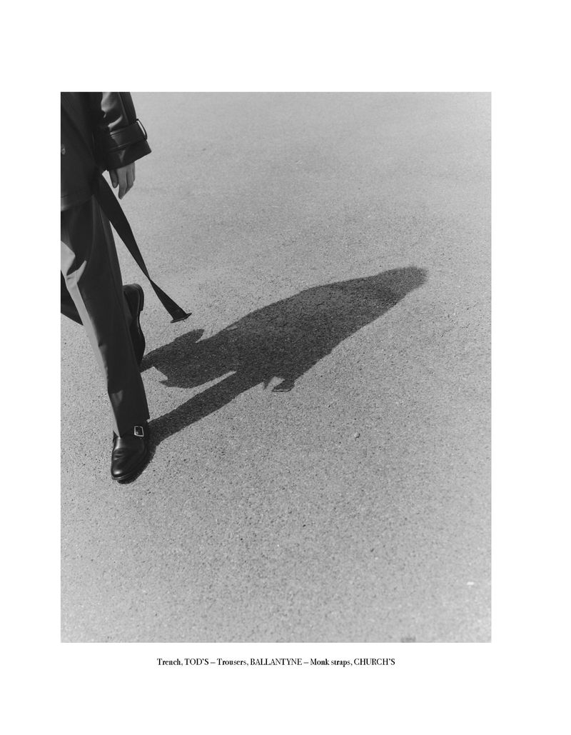 A black and white fashion shot of a person in a coat and black shoes walking on pavement, with their shadow cast in front of them.