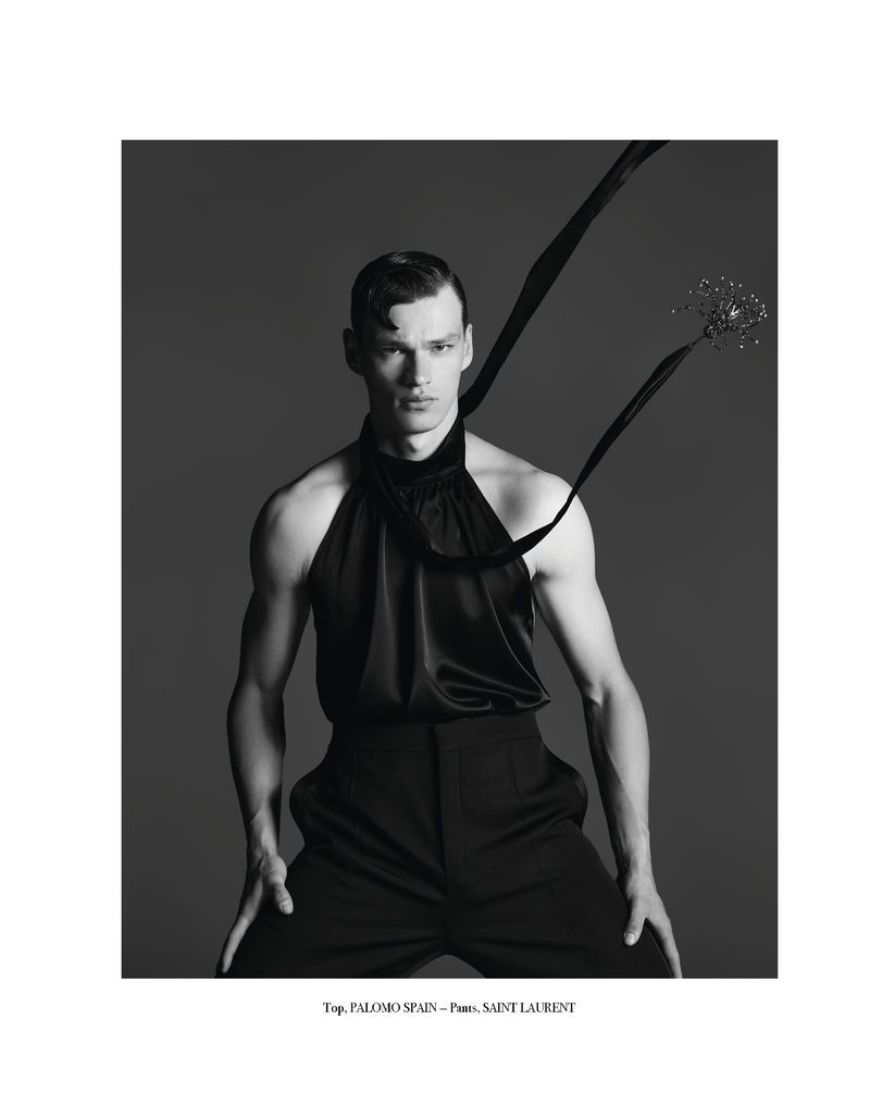 Black and white studio shot of a male model wearing a halter top and black pants, posing against a dark gray backdrop with ribbon detail.