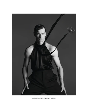 Black and white studio shot of a male model wearing a halter top and black pants, posing against a dark gray backdrop with ribbon detail.