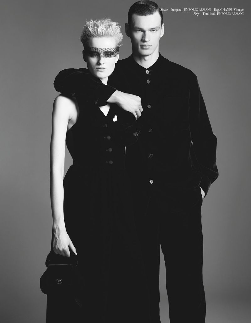 Black and white fashion shot of a man in a black suit and woman in a sleeveless dress with ornate eye mask, both looking forward.