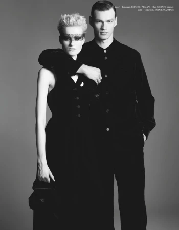 Black and white fashion shot of a man in a black suit and woman in a sleeveless dress with ornate eye mask, both looking forward.