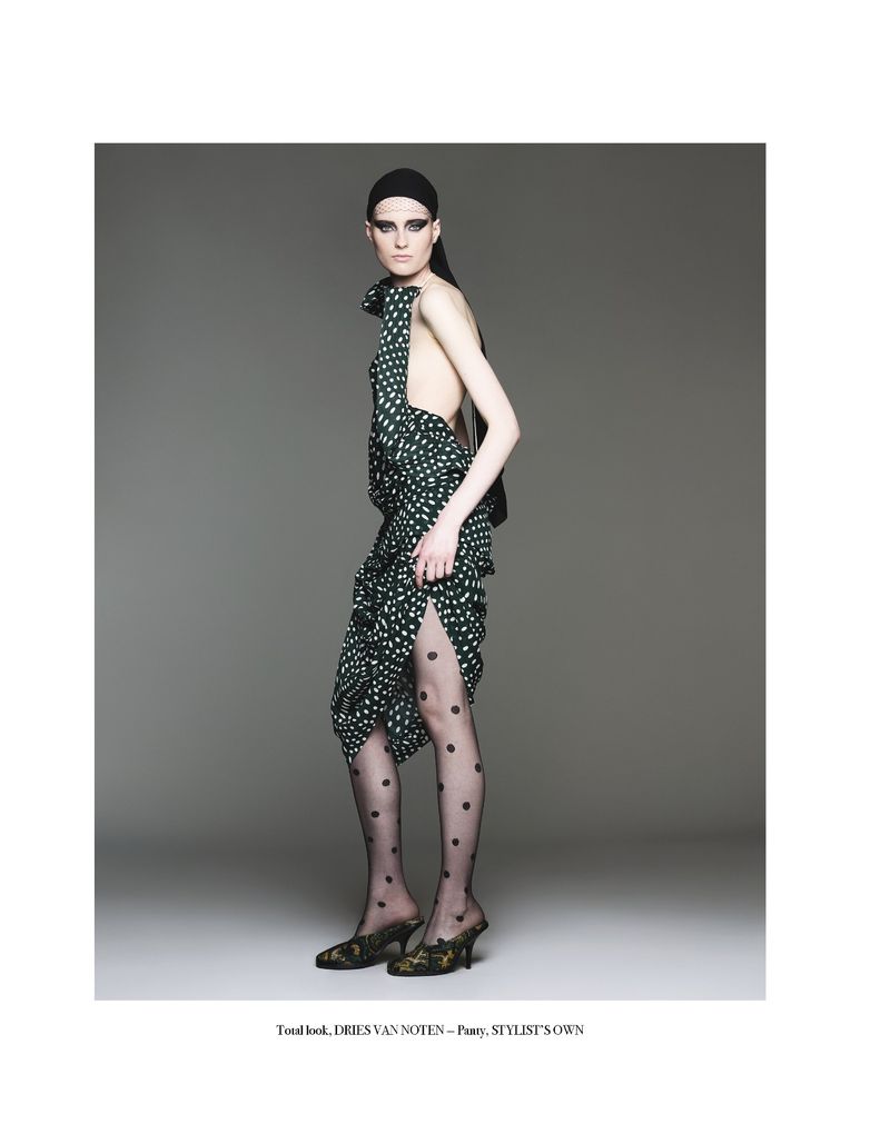Fashion photo of an adult woman wearing a green and white polka-dot dress, black polka-dot tights, and heeled shoes against a gray background.
