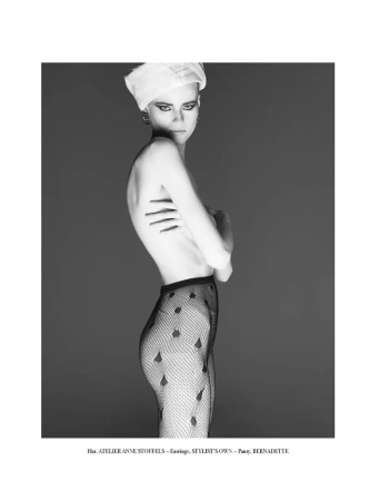 Monochrome fashion shot of a slender woman with a white head wrap and patterned tights posing in front of a gray background.