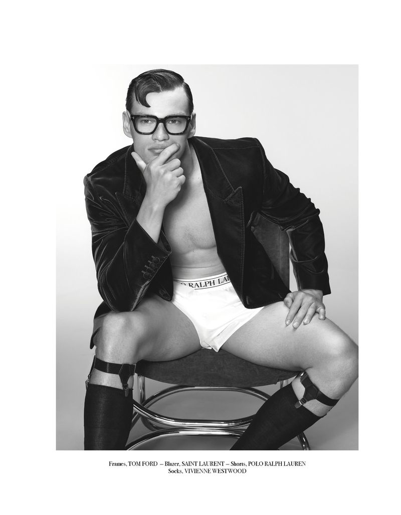 Black and white studio shot of a man in eyeglasses, briefs, gartered stockings, and a velvet jacket sitting on a stool with hand to chin.