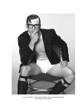 Black and white studio shot of a man in eyeglasses, briefs, gartered stockings, and a velvet jacket sitting on a stool with hand to chin.