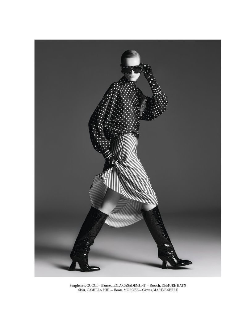 A woman in a black and white polka dot blouse, striped skirt, sunglasses, and boots poses against a gray backdrop in a studio setting.