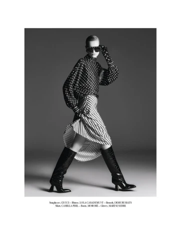 A woman in a black and white polka dot blouse, striped skirt, sunglasses, and boots poses against a gray backdrop in a studio setting.