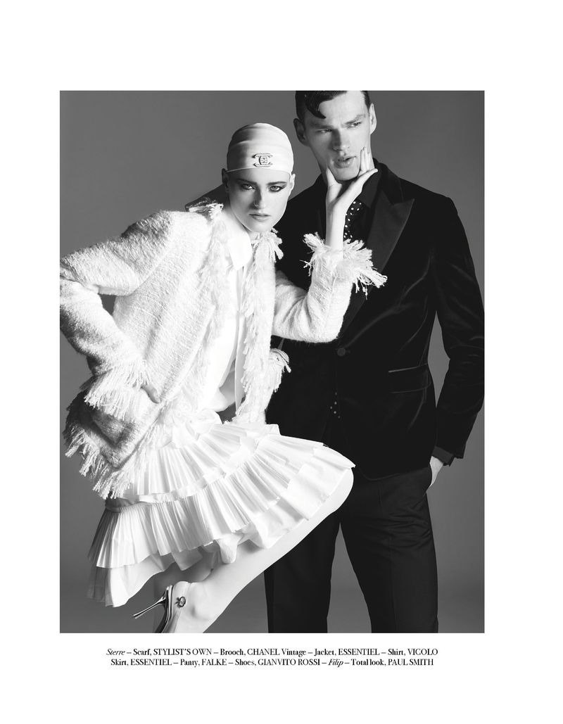 Editorial fashion shot with a female model in a Chanel headscarf, white jacket and skirt, and a male model in a black velvet suit jacket.
