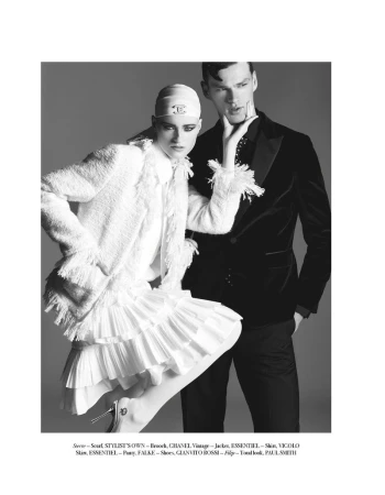Editorial fashion shot with a female model in a Chanel headscarf, white jacket and skirt, and a male model in a black velvet suit jacket.