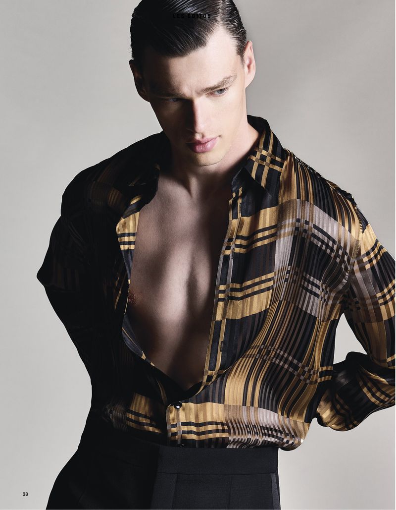 Man poses in unbuttoned black and gold patterned shirt and black pants against a grey background in a studio fashion shot.