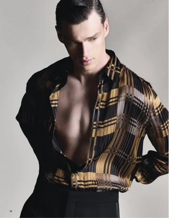 Man poses in unbuttoned black and gold patterned shirt and black pants against a grey background in a studio fashion shot.