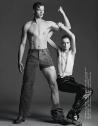 Black and white studio portrait of a shirtless male model standing and a seated female model posing against a grey backdrop.