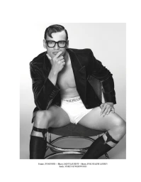 Model Filip Hrivnak seated wearing dark-rimmed glasses, an open blazer, white briefs, and sock garters against a studio sweep.