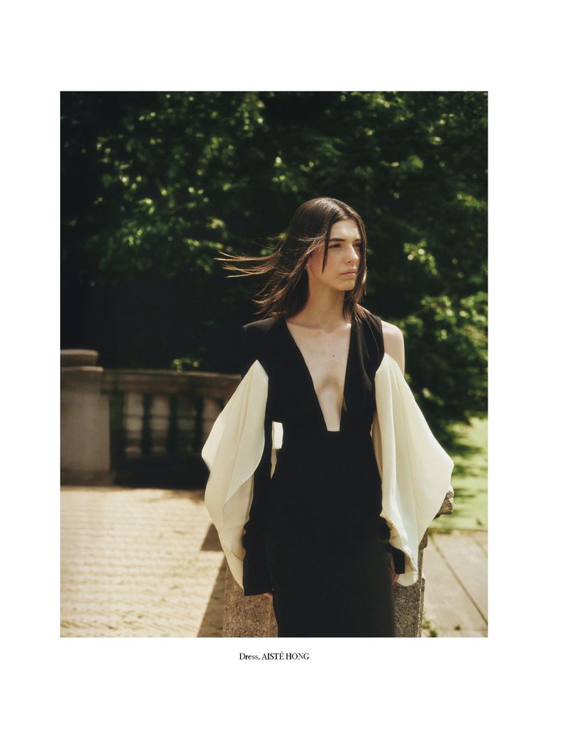 Fashion editorial shot of a brunette woman in a black dress with flowing white sleeves posing outdoors on a stone path surrounded by trees.