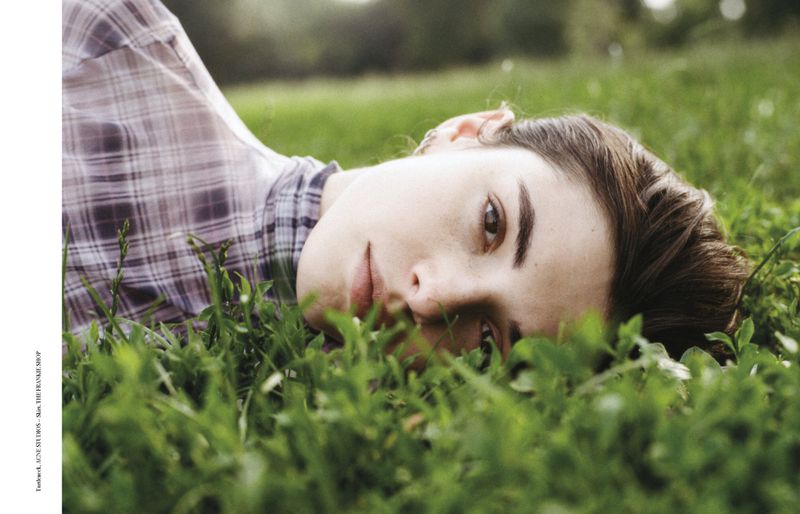 Eye-level close-up of a brunette woman lying on grass outdoors, wearing a plaid shirt, with blurred foliage background in soft, natural light.