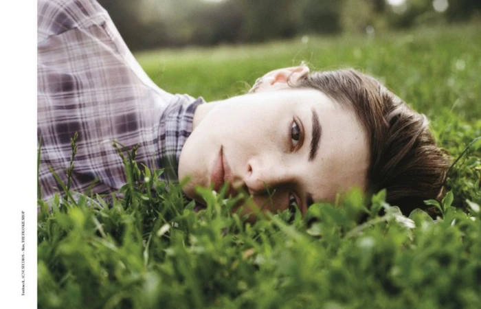 Eye-level close-up of a brunette woman lying on grass outdoors, wearing a plaid shirt, with blurred foliage background in soft, natural light.