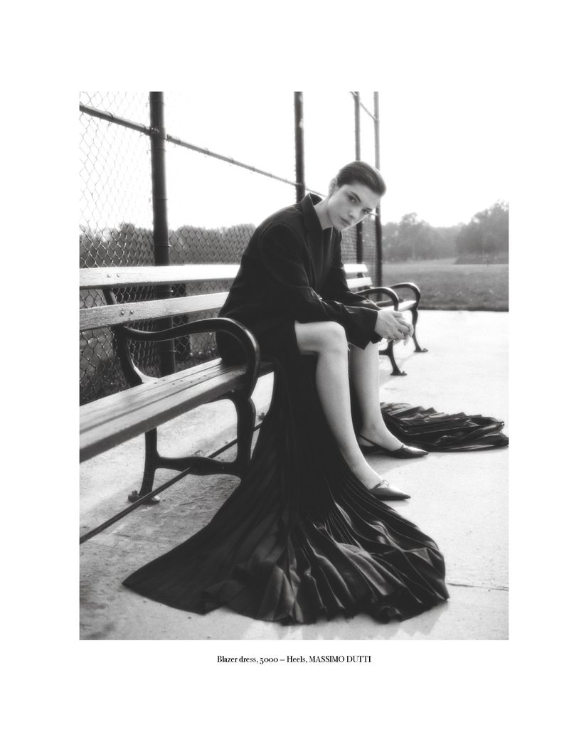 Monochrome fashion shot: woman poses on park bench in a dark dress and jacket, casting a gaze at the camera in soft daylight.