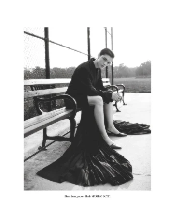 Monochrome fashion shot: woman poses on park bench in a dark dress and jacket, casting a gaze at the camera in soft daylight.