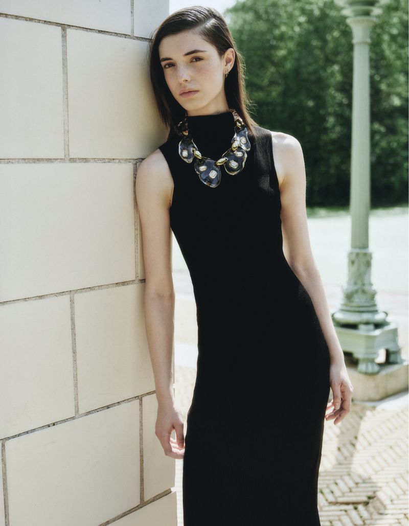 A young woman with dark hair in a black dress leans against a white wall, wearing a statement necklace, in a natural outdoor setting.