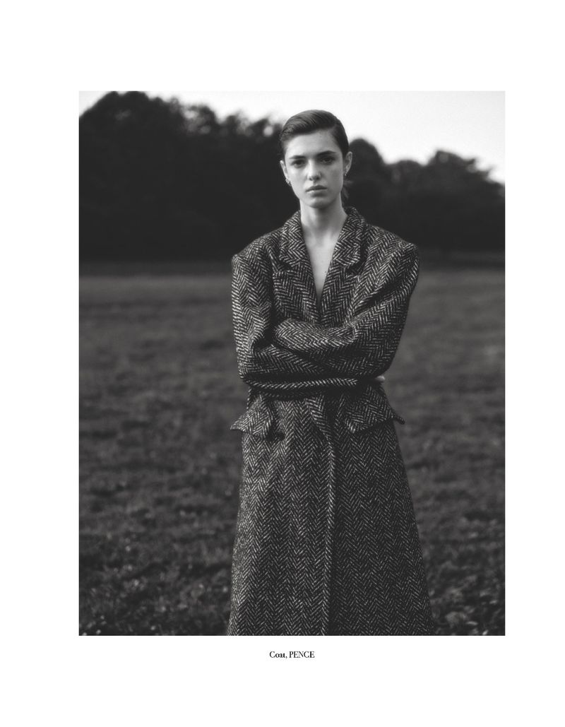 Fashionable woman with arms crossed poses in a herringbone coat outdoors in a grassy field surrounded by trees in a black and white portrait.