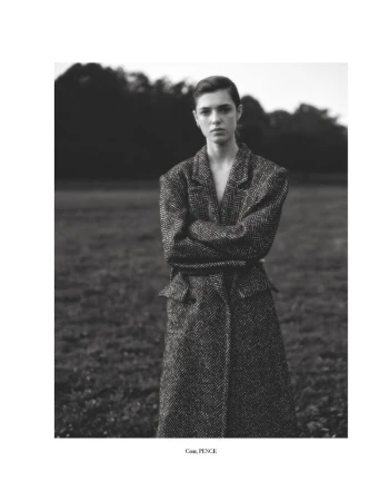 Fashionable woman with arms crossed poses in a herringbone coat outdoors in a grassy field surrounded by trees in a black and white portrait.