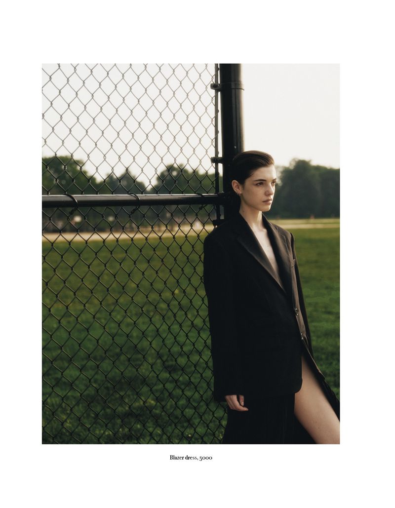 A woman with short dark hair stands next to a fence on a grassy field, wearing an oversized dark blazer, in an outdoor setting.