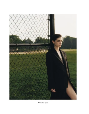 A woman with short dark hair stands next to a fence on a grassy field, wearing an oversized dark blazer, in an outdoor setting.