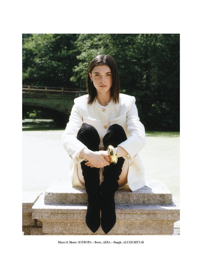 An adult woman sits on a concrete ledge in front of a park, wearing a cream-colored jacket, black boots, and a gold bracelet.