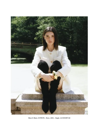 An adult woman sits on a concrete ledge in front of a park, wearing a cream-colored jacket, black boots, and a gold bracelet.
