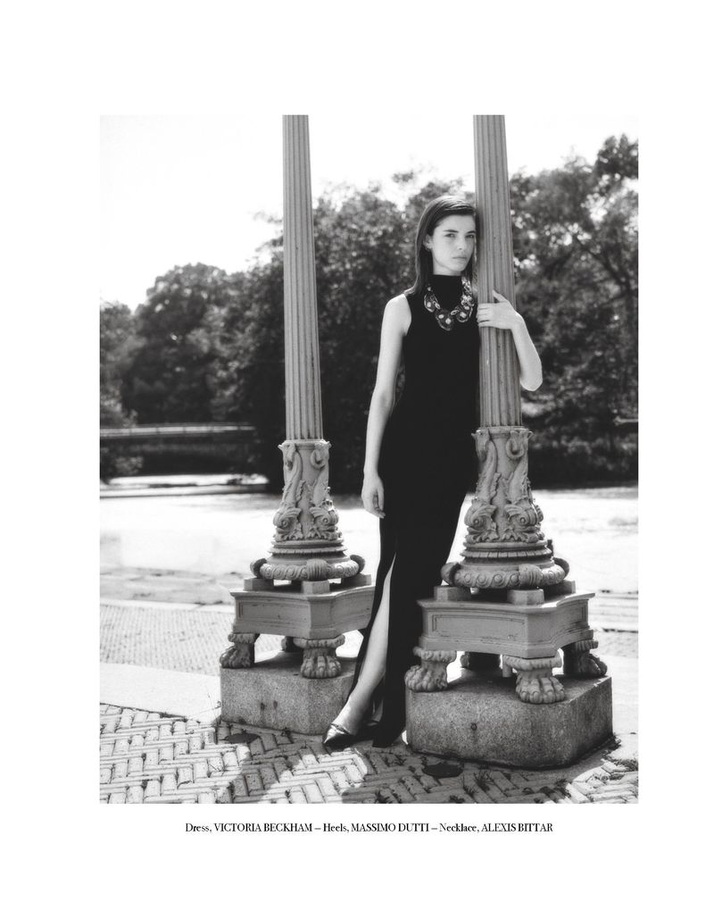 A stylish woman in a black dress and necklace poses near two ornate pillars in a black and white fashion photograph taken outdoors.