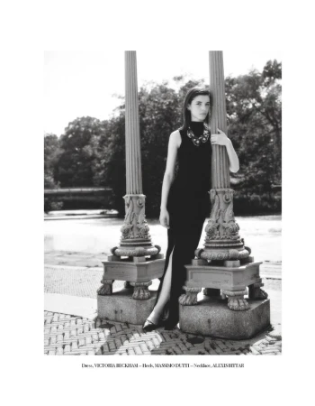 A stylish woman in a black dress and necklace poses near two ornate pillars in a black and white fashion photograph taken outdoors.