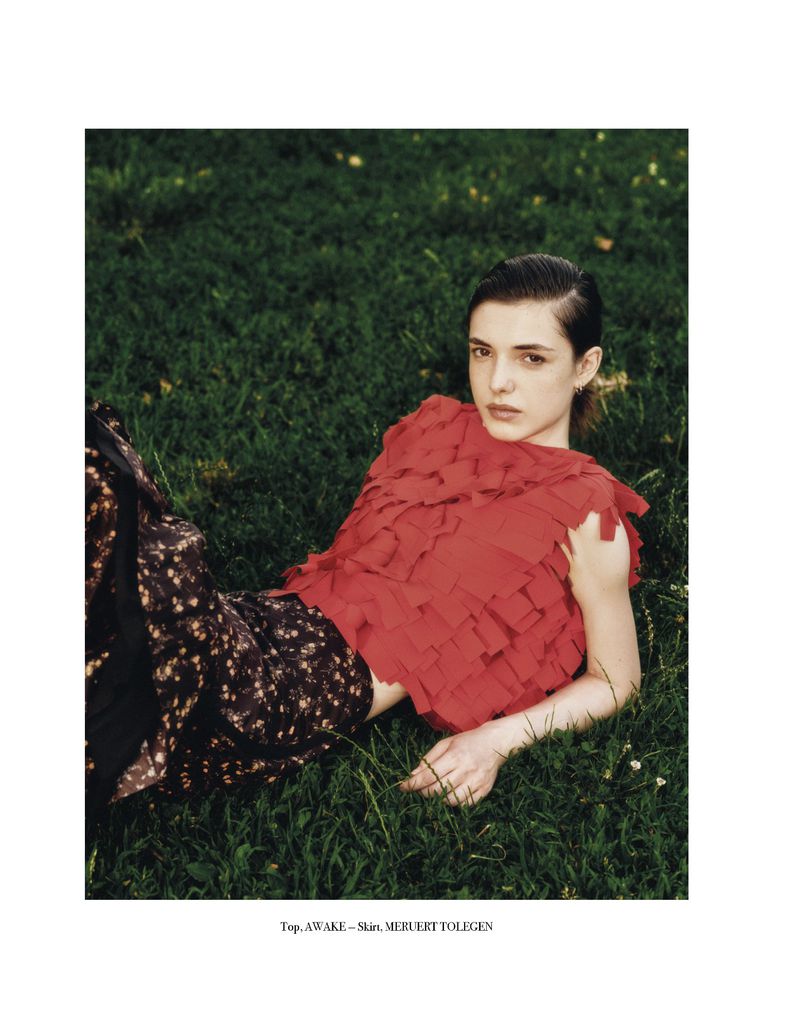 Fashion editorial shot of a young woman with dark hair in a red, frilly top and patterned pants, lying in green grass outdoors.