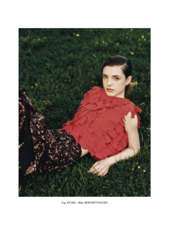 Fashion editorial shot of a young woman with dark hair in a red, frilly top and patterned pants, lying in green grass outdoors.