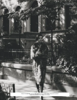 Black and white editorial photo of model Carla Rey wearing a textured fur coat on a sidewalk before brownstone residential buildings.