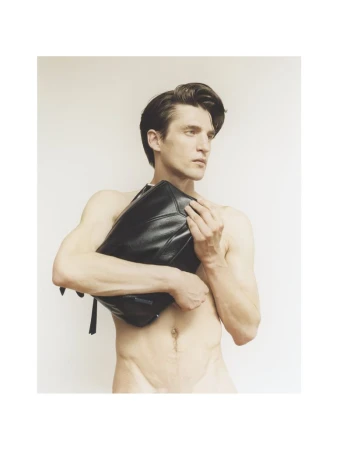A nude male model poses in a studio with a shiny black clutch bag held to his chest, against a neutral studio backdrop.
