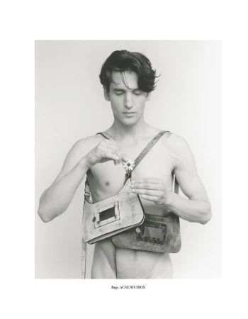 Black and white studio shot of an adult male holding a daisy, wearing two fashion purses from Acne Studios, minimalist neutral backdrop.