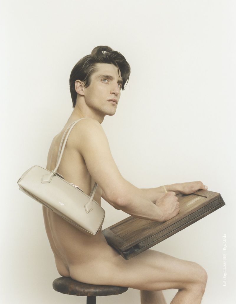 Nude man sits on a stool holding a drawing board, wearing a beige purse slung over his shoulder, in a fashion photography studio setting.