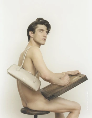 Nude man sits on a stool holding a drawing board, wearing a beige purse slung over his shoulder, in a fashion photography studio setting.