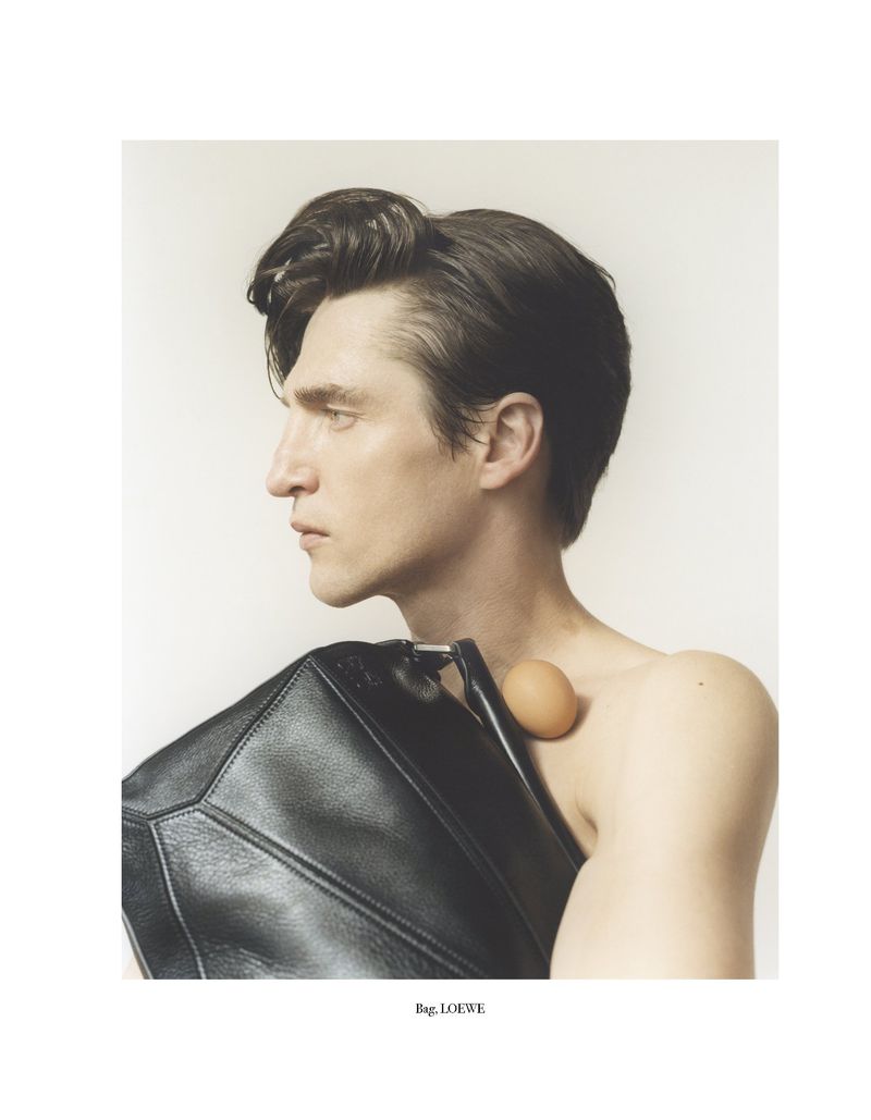 Fashion portrait of a bare-chested adult male with black hair holding a black leather handbag and an egg on his shoulder in a studio setting.