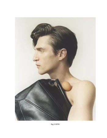 Fashion portrait of a bare-chested adult male with black hair holding a black leather handbag and an egg on his shoulder in a studio setting.