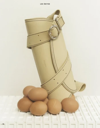 Beige cylindrical purse with black stitching detail and multiple straps around brown eggs displayed on a white slatted surface.