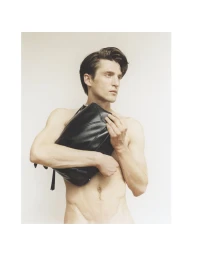 Model Anatol Modzelewski holding a black leather bag across his chest against an off-white seamless backdrop.