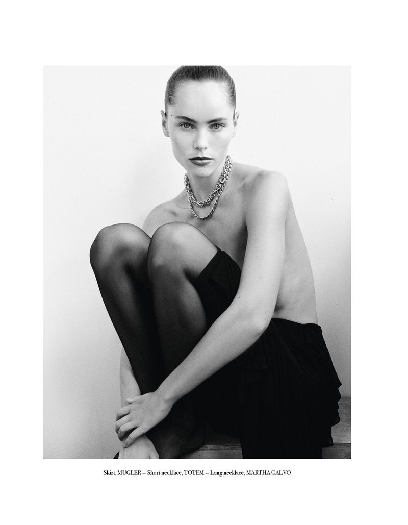 A woman with dark hair poses seated with a chain necklace, and black tights in a studio shot against a light background.