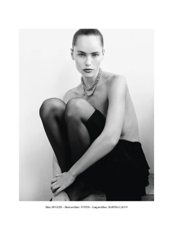 A woman with dark hair poses seated with a chain necklace, and black tights in a studio shot against a light background.